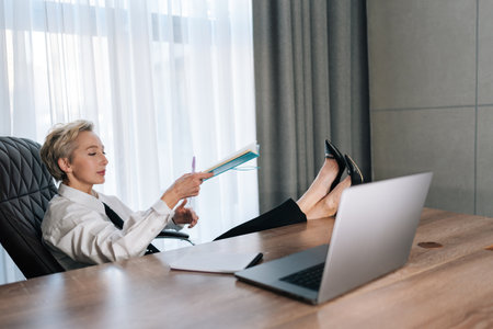 Relaxed middle-aged female CEO putting feet on desk with laptop. Confident successful business woman thinking about career growth relaxing in cabinet. Concept relaxation at workplaceの写真素材