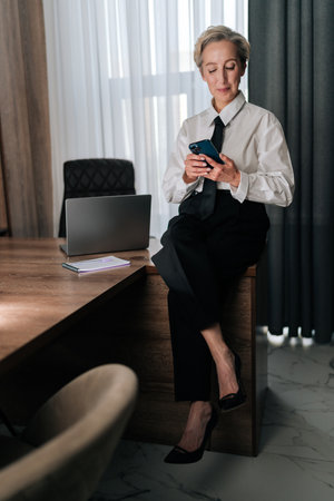 Vertical portrait of focused middle-aged business woman sitting on office table, looking at cellphone screen, making call by business, search information on internet distracted from work.の写真素材