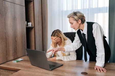 Aggressive middle-aged woman boss scolding female employee for bad result at workplace. Dissatisfied executive pointing at laptop reprimanding incompetent intern for ineffective poor performanceの写真素材
