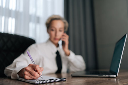 Selective focus portrait of busy middle-aged business woman multitasking in office, working online with laptop, talking smartphone, writing documents with pen sitting at office desk by window.の写真素材