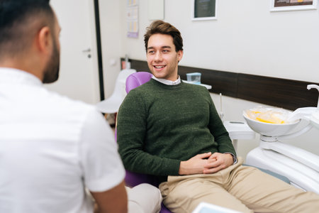 Portrait of cheerful bearded young man patient attentively listening unrecognizable male dentist discusses treatment plan sitting on dental chair in dentistry clinic. Concept of dentistry, teeth care.の写真素材