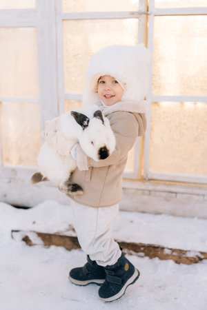 Vertical portrait of happy little child boy in warm clothes hugging white funny rabbit standing by farm house on sunny winter day, looking at camera. Concept of excursion to eco-farm, life in village.の写真素材