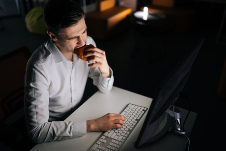 High-angle view of freelancer male eating burger working on desktop computer sitting on desk at office workplace late night in dark room. Businessman eating fast food at laptop and reading email.の写真素材
