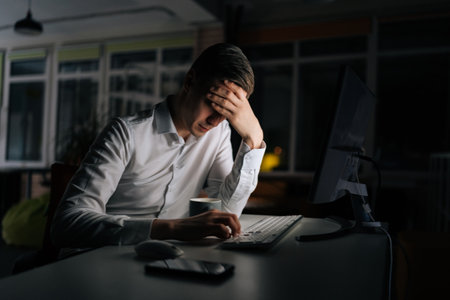 Exhausted businessman working in evening office alone. Tired anxious manager losing failure working on computer at late night. Tired worried business man at workplace in office holding head on hands.の写真素材