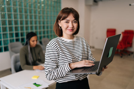 Portrait of friendly young businesswoman in casual clothes holding laptop in hand standing in office smiling looking at camera. Diverse business team discussing project sitting at desk on backgroundの写真素材