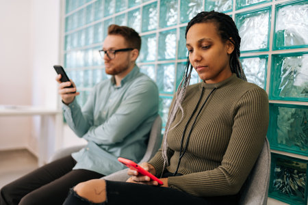 Two diverse young business people, hiring, using mobile phones in waiting room for networking, social media or communication at office. Group of employee workers wait in line with smartphoneの写真素材