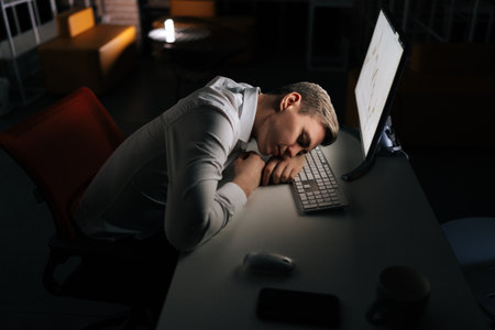 Tired male trader asleep on computer desk late at night alone. Exhausted yougng businessman sleeping with head on keyboard while working overnight on computer. Concept of overwork and deadline.の写真素材