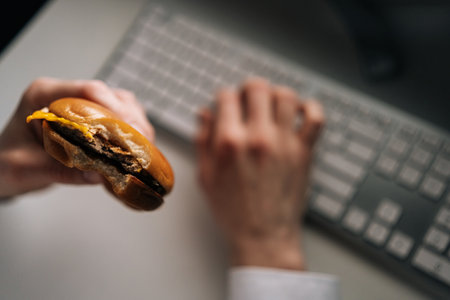 Detail cropped shot of unrecognizable freelancer male holding eating burger working in office at night. Closeup of male employee sitting on table, feeling hungry, having dinner in corporate workplaceの写真素材