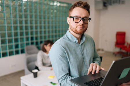 Portrait of confident startup businessman in casual clothes and glasses holding laptop in hand...