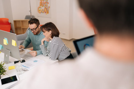 Rear view of male mentor and female intern sitting at desk with laptop doing paperwork together discussing project financial report, at creative open space. Corporate business collaboration concept.の写真素材