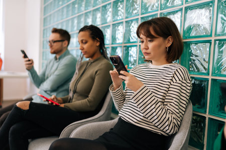 Serious multicultural young business people sitting in row using smartphones, waiting for job interview, sitting in queue line row in chairs in modern office waiting room.の写真素材