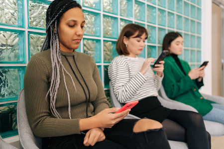 Carefree multiethnic casual young people sitting in lobby of civil service building browsing web pages in Internet on smartphone while waiting to meet with representative.の写真素材