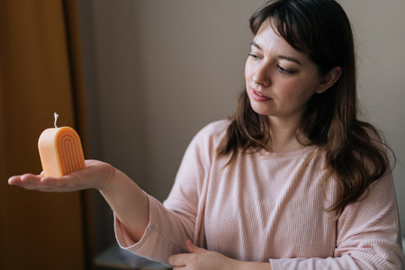 Portrait of pretty female artisan craftperson holding in hand finished candle after create. Process of making handmade natural scented candle. Creative occupation of making trendy DIY candles.の写真素材