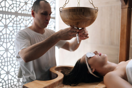 Portrait of spa mater gently guiding oil stream on female client forehead during extraordinary Shirodhara treatment, Ayurvedic healing technique in wellness salon. Concept of body care, rehabilitationの写真素材