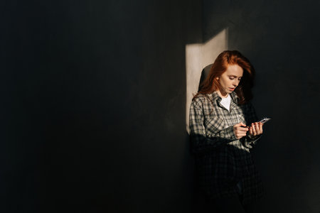 Beautiful young woman holding using mobile phone at shadow background in dark living room, looking to smartphone screen, enjoying doing online ecommerce shopping in mobile apps indoors.の写真素材