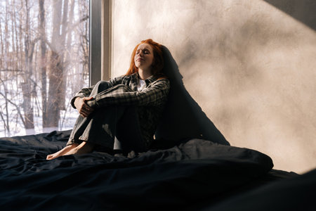 Portrait of sad young woman sitting by window on sunny day on windowsill with winter nature background, feels upset, having melancholic mood, thinking about life troubles. Loneliness concept.の写真素材