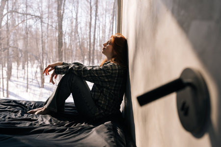 Side view of crying young woman sitting by window on sunny day on windowsill with winter nature background, feels upset, having melancholic mood, thinking about life troubles. Loneliness concept.の写真素材