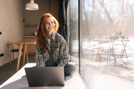 Portrait of happy freelancer female remote working on laptop sitting on wide windowsill with nature view on sunny day. Cheerful young student typing on laptop keyboard, using online app at home.の写真素材