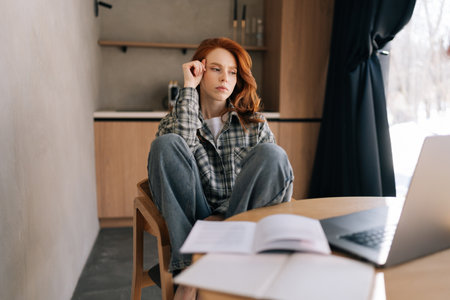Millennial redhead businesswoman working from home office using laptop, staring at device screen looking pensive and thoughtful, search solution, prepare report or research, doing freelance job.の写真素材