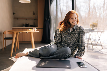 Portrait of charming redhead young woman sitting by window on sunny day relaxing at home looking at camera. Smiling calm female chilling at house, dreaming, enjoying peace of mind.の写真素材