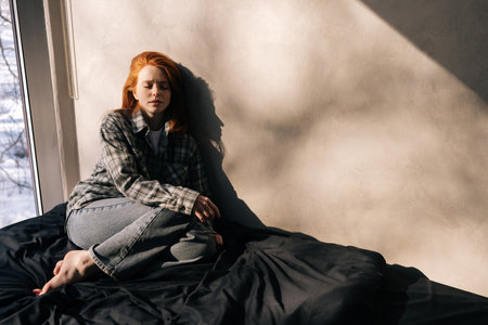 Portrait of depressed young woman sitting on sunny day on floor by window alone, having melancholic mood, troubled with loneliness, upset about solitude mental problem, regret bad mistake concept.の写真素材