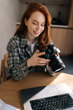 Vertical portrait of young female photographer holding modern digital camera looking at screen choosing photos for editing while sitting in front of laptop computer at home office workplace.の写真素材