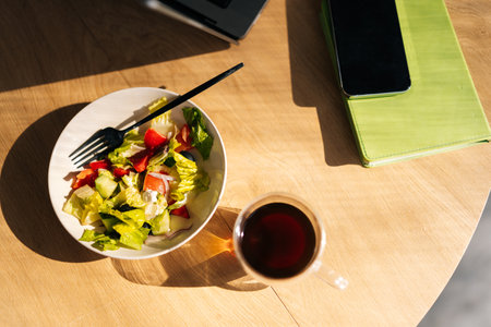Top view of fresh vegetable salad, laptop, smartphone and glass cup of coffee on wooden table at workplace, nobody. Business lunch. concept of food delivery, lunch in the office.の写真素材