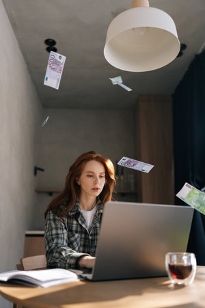 Vertical shot of money cash dollar rain falling on pretty young businesswoman sitting at home desk...