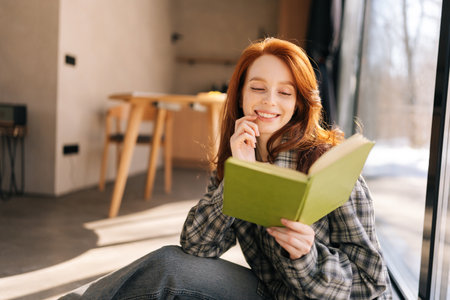 Portrait of cheerful young redhead woman holding reading book sitting by window on sunny day, smiling enjoying leisure activity, having carefree weekend with favorite literature and home pastime.の写真素材