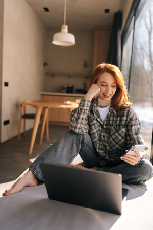 Vertical shot of positive young woman typing on cellphone at home office workplace, sitting by window on sunny day, working at laptop, getting positive, thoughtful, looking to smartphone screenの写真素材