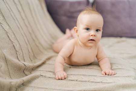 Portrait of cute little baby girl lying on stomach on bed, interestingly looking at camera with open...