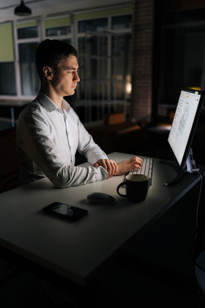 Vertical shot of serious young office worker male working at workplace on computer at late night in dark room. Successful entrepreneur checking finances on internet sitting at desk at evening.の写真素材