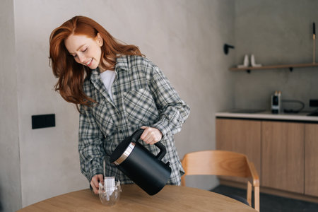 Attractive smiling young woman making coffee with drip coffee bag in glass transparent cup standing by table in morning. Process preparing fresh brewed drip coffee in cup with paper drip bag.の写真素材
