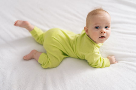 High-angle view of adorable newborn baby girl wearing yellow cotton bodysuit lying on stomach on white sheet. Newborn child relaxing in bed in children nursery, looking at camera.の写真素材