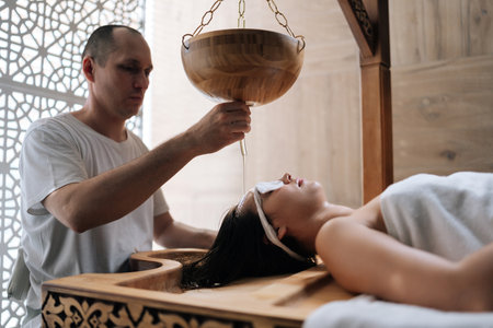 Relaxed young woman peaceful resting while stream of oil covering her forehead. Diligent experienced master actively adding oil stream on forehead of peaceful client. Concept of luxury spa procedure.の写真素材