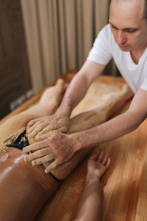 Vertical shot of male massage therapist scrubbing massaging tanned buttocks with warm sand of female client lying on table in wellness center. Lymphatic drainage massage of legs in spa salon.の写真素材