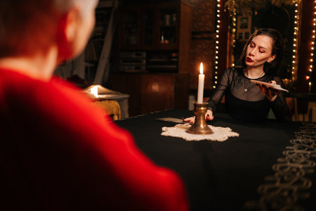 Closeup view from shoulder of unrecognizable female client getting divination session with tarot cards at dark esoteric room. Mysterious seer talking to client at table with tarot card layout.の写真素材