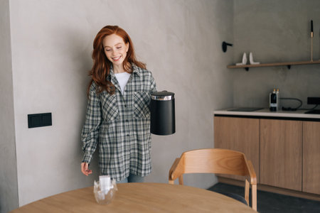 Portrait of happy smiling young woman making coffee with drip coffee bag in glass transparent cup standing by wooden table. Process preparing fresh brewed drip coffee in cup with paper drip bag.の写真素材