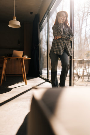 Low-angle view of young woman having pleasant phone call, standing relaxing near window in living room on sunny day. Portrait of redhead female talking on smartphone and looking out window at home.の写真素材