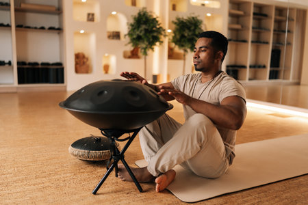 Portrait of relaxed African man playing traditional ethnic music using glucophone sitting on floor in lotus pose. Serene black male in casual clothes drumming rhythm on tank drum by hands.の写真素材