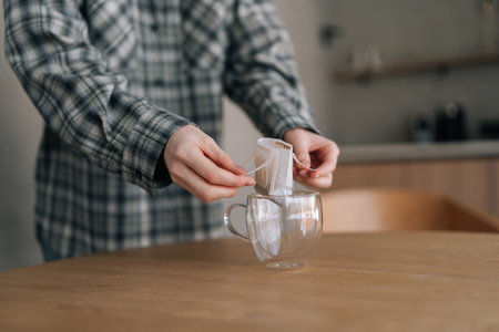 Close-up cropped shot of unrecognizable woman putting bag of drip coffee in glass cup at table in...
