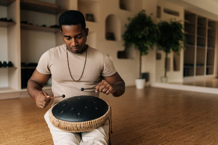 Front view of African man playing ethnic music on tank drum using drumsticks sitting on floor in...