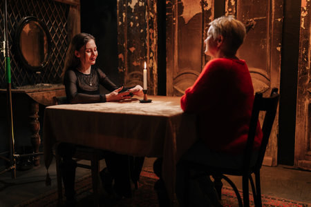 Side view from of middle-aged female client getting divination session with tarot cards in dark esoteric room. Mysterious sorceress talking to client at table with tarot card layout.の写真素材
