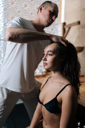 Closeup vertical portrait of relaxed young female client with dark hair and bare shoulder getting head massage by professional masseur in wellness center. Concept of rest with spa treatment.の写真素材