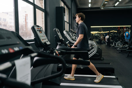 Full length side shot of athletic man walking on treadmill preforming warming-up run routine exercise in gym. Sporty male walking on running track, working on health and fitness at sport club.の写真素材