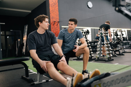 Young tired man in conversation with personal trainer in gym after workout training on rowing machine. Sweaty male taking break after workout exercise. Exhausted people talking to each others.の写真素材