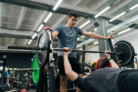 Low-angle view of experienced fitness instructor helping beginner sportsman doing barbell bench press exercise during personal workout in gym. Male doing barbell bench press under couch supervisionの写真素材