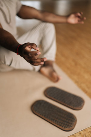 Vertical cropped shot of unrecognizable black male meditating in lotus pose at home sitting on yoga mat by sadhu board. African man prepares for practice. Concept of mindfulness, tranquility, harmony.の写真素材