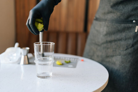Cropped shot of unrecognizable bartender male in black gloves tosses kiwi slices into glass to make deliciously refreshing cocktail in summer cafe. Concept of party time, holidays, celebration mood.の写真素材