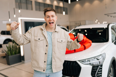 Joyful customer male standing by new car with huge red bow in dealership, looking at camera, showing positive gesture. Buyer man feeling happy after successful purchase. Concept of buying new auto.の写真素材